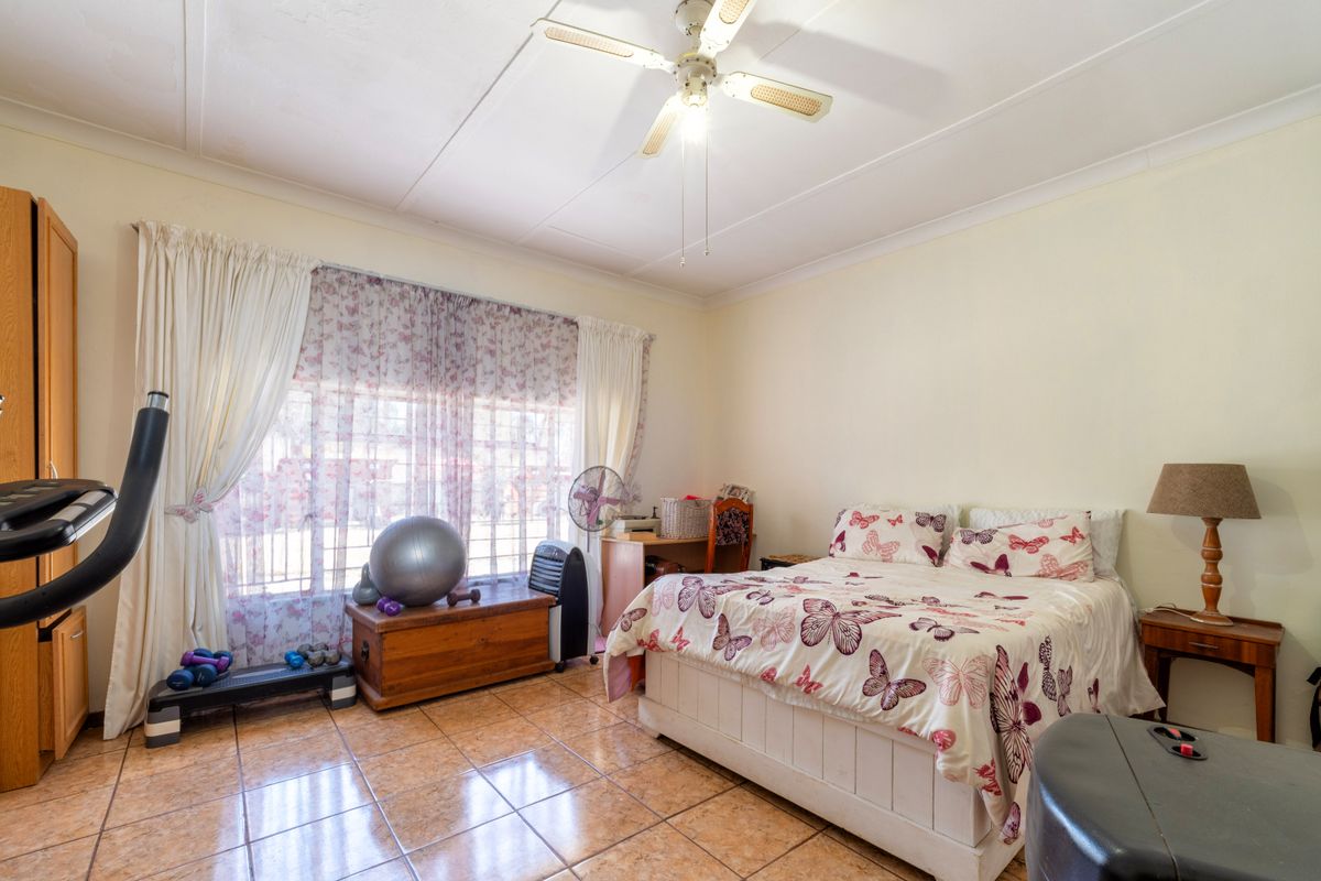 A spacious bedroom featuring tiled flooring, a ceiling fan, and a built-in cupboard, with large windows secured by burglar bars.