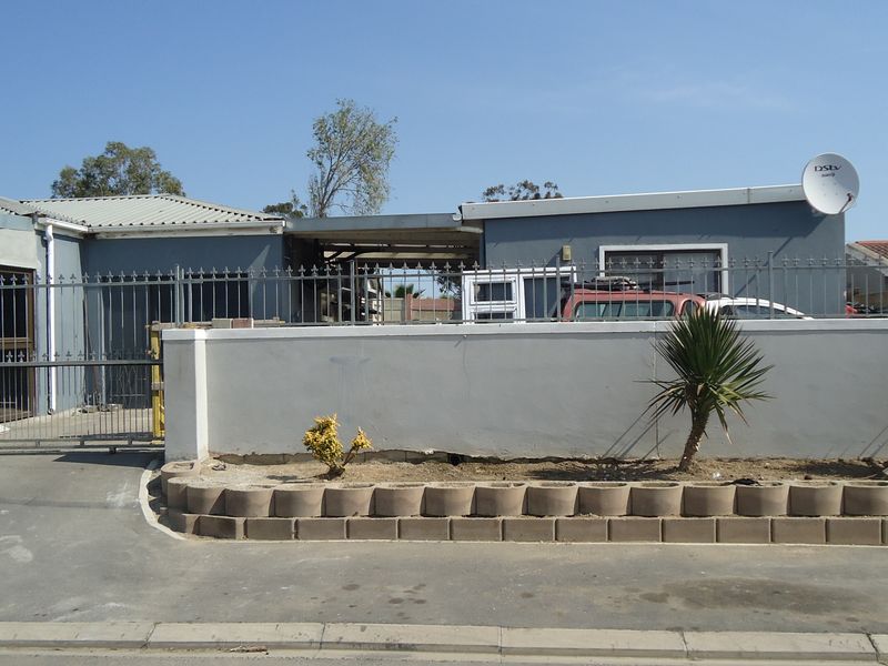 Front exterior view of the house featuring a metal sliding gate, perimeter wall, and concrete driveway with visible areas of unfinished plaster.