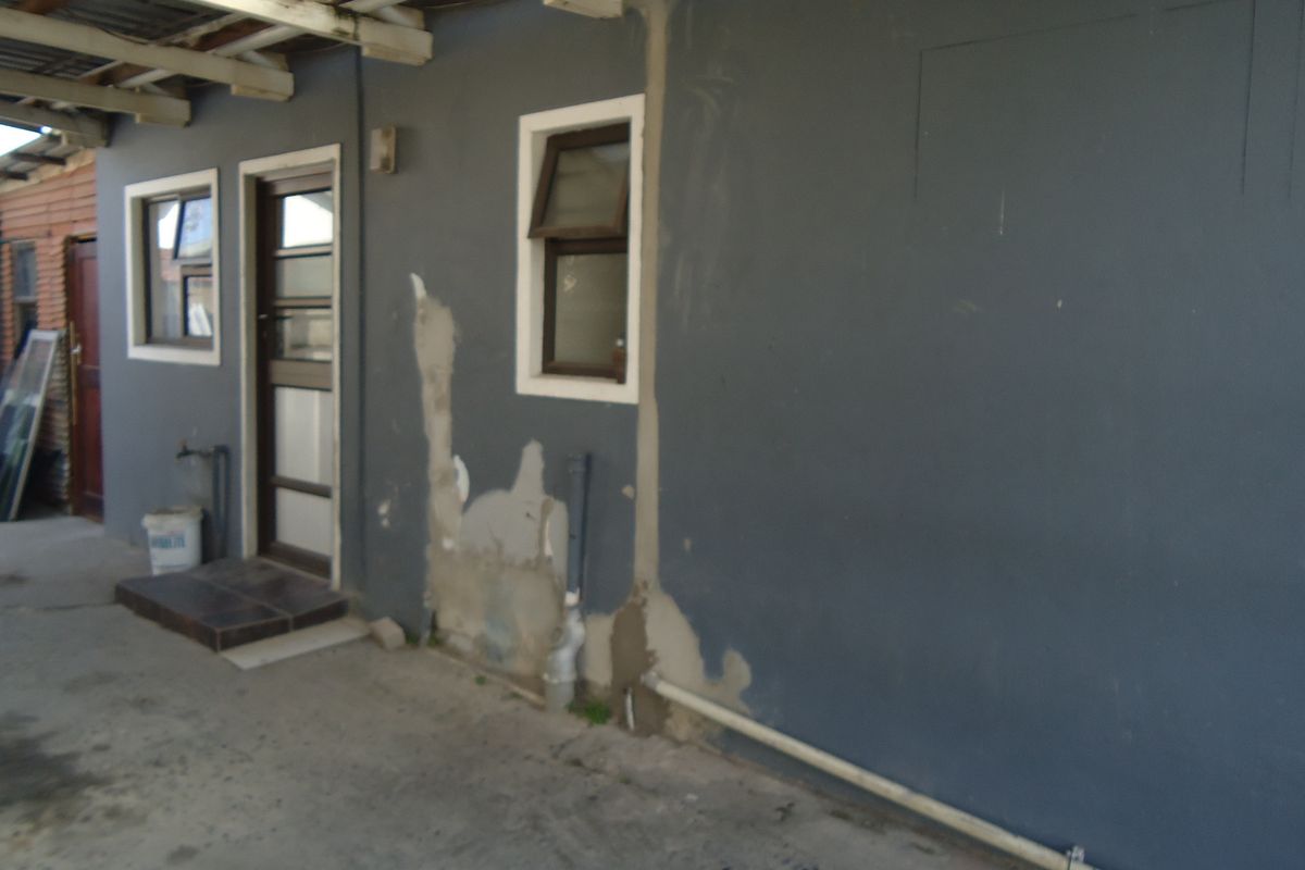 Rear exterior area featuring a concrete patio, external access door, and casement windows, currently requiring maintenance to the wall finishes and paintwork.