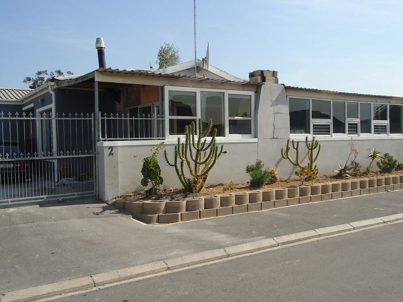 Front exterior view of a single-story house featuring a metal security gate, concrete retaining wall, and large aluminium-framed windows.
