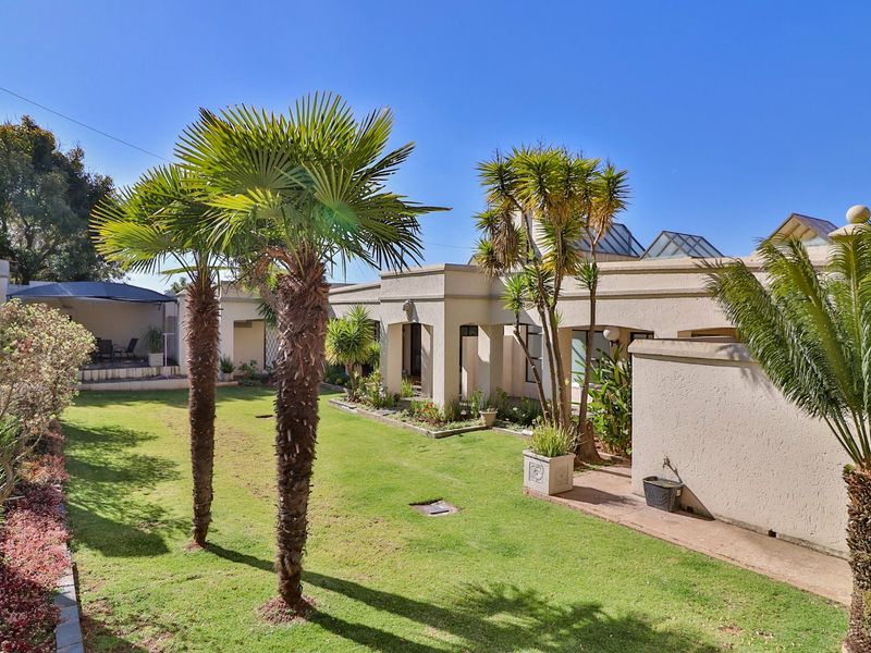 A well-maintained rear garden featuring a manicured lawn, mature palm trees, and a paved walkway leading to a covered patio area.