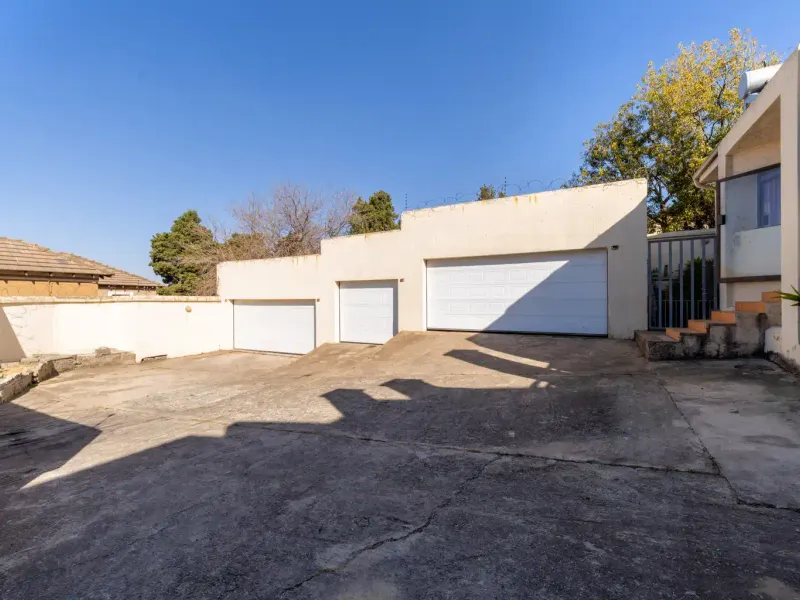 Exterior view of a property featuring a triple garage, concrete driveway, and security fencing, showing signs of wear on the driveway and exterior walls.
