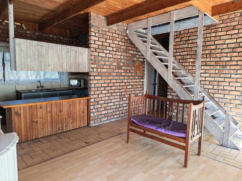 Open plan living area featuring exposed brick walls, timber ceiling beams, a wooden staircase, and a functional kitchen space with tiled and laminate flooring.