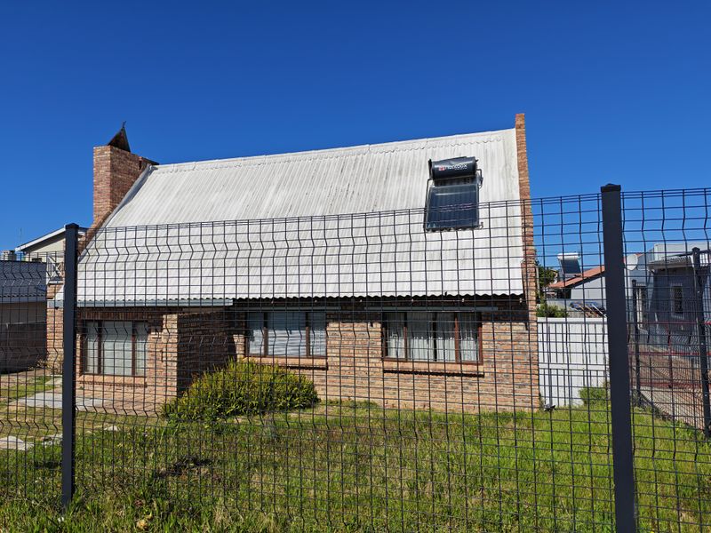 A single-story face brick house featuring a corrugated metal roof, a solar water heating system, and secure wire mesh perimeter fencing.