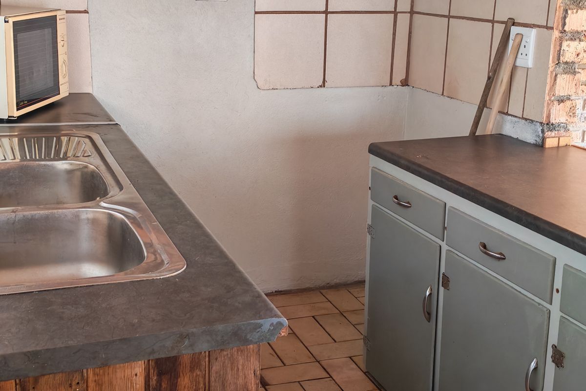 A dated kitchen area featuring a double stainless steel sink, laminate countertops, tiled backsplash, and original built-in cabinetry with tiled flooring.