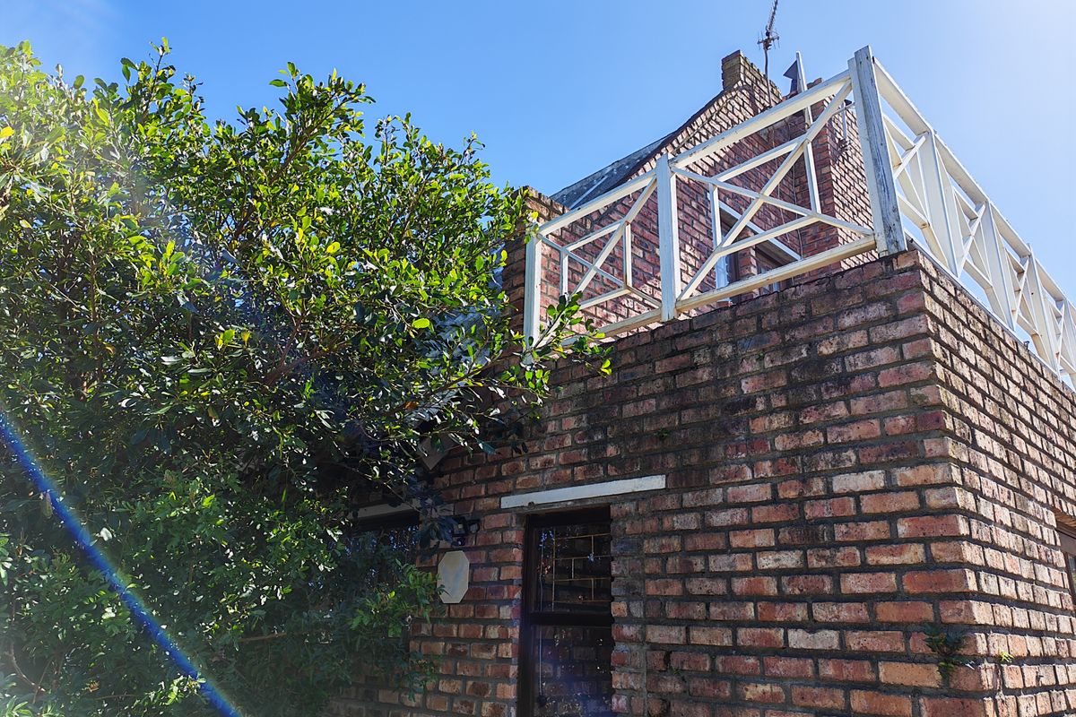 Exterior view of a brick house featuring an upper-level balcony with a white timber balustrade and mature garden foliage.