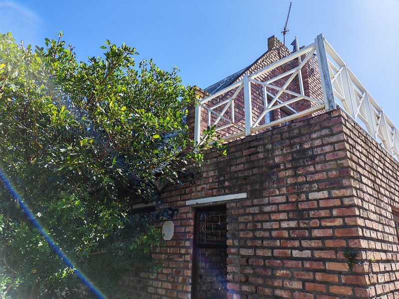 Exterior view of a brick house featuring an upper-level balcony with a white timber balustrade and mature garden foliage.