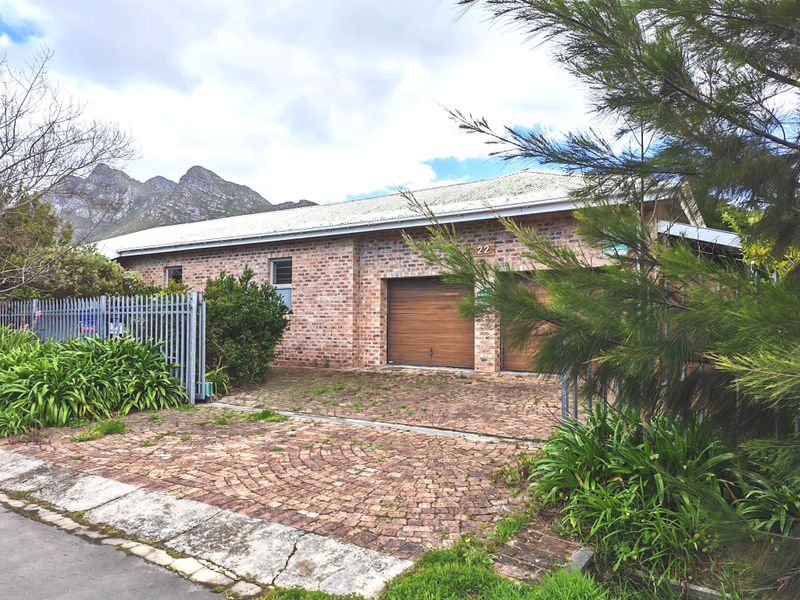 Front exterior view of a brick house featuring a double garage, paved driveway, and secure metal fencing against a mountain backdrop.