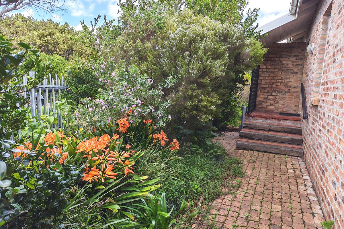 A rear exterior view featuring a brick-paved pathway, timber steps leading to the building entrance, and established garden landscaping.