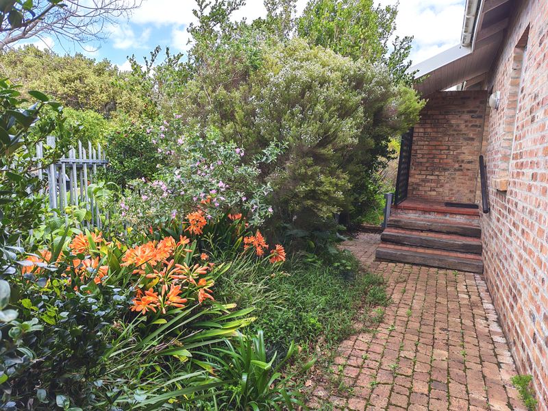A rear exterior view featuring a brick-paved pathway, timber steps leading to the building entrance, and established garden landscaping.