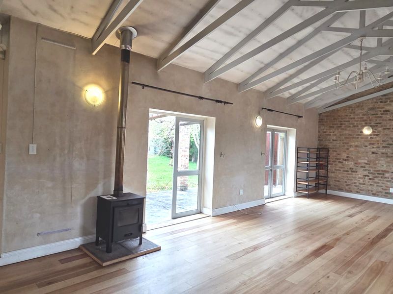 Spacious living area featuring exposed timber beams, a wood-burning fireplace, hardwood floors, and an exposed brick feature wall with direct access to the garden.