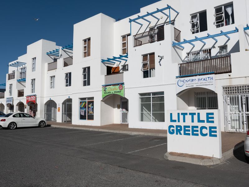 Exterior view of the Little Greece apartment complex featuring a paved parking area, ground floor commercial units, and upper-level residential balconies.