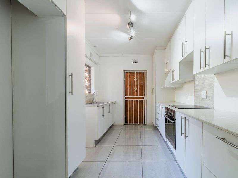 A clean, modern galley-style kitchen featuring white cabinetry, stone countertops, integrated oven and hob, and tiled flooring.