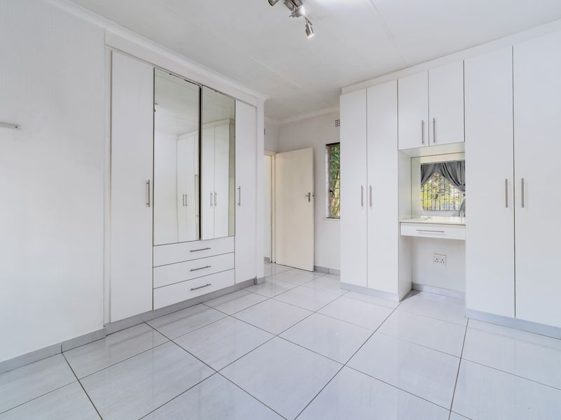 A bright, empty bedroom featuring extensive white built-in cabinetry, a mirrored wardrobe, a built-in vanity station, and clean tiled flooring.