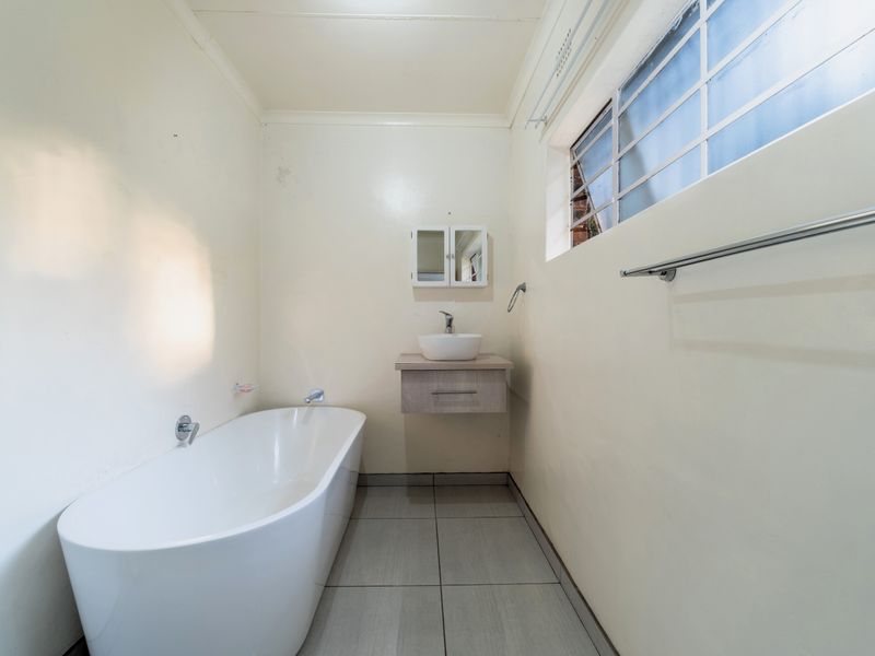 A clean, minimalist bathroom featuring a modern freestanding bathtub, a wall-mounted vanity with a vessel sink, and neutral tiled flooring.