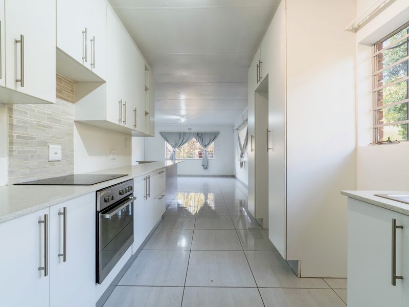 A bright, modern galley-style kitchen featuring white cabinetry, integrated appliances, tiled flooring, and a neutral backsplash, leading into an open-plan living area.