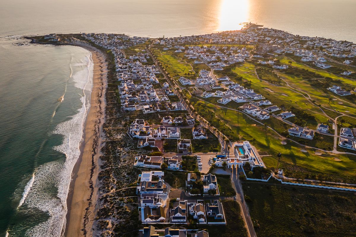 An expansive aerial view of a coastal residential estate featuring a golf course, beach access, and well-maintained properties at sunset.