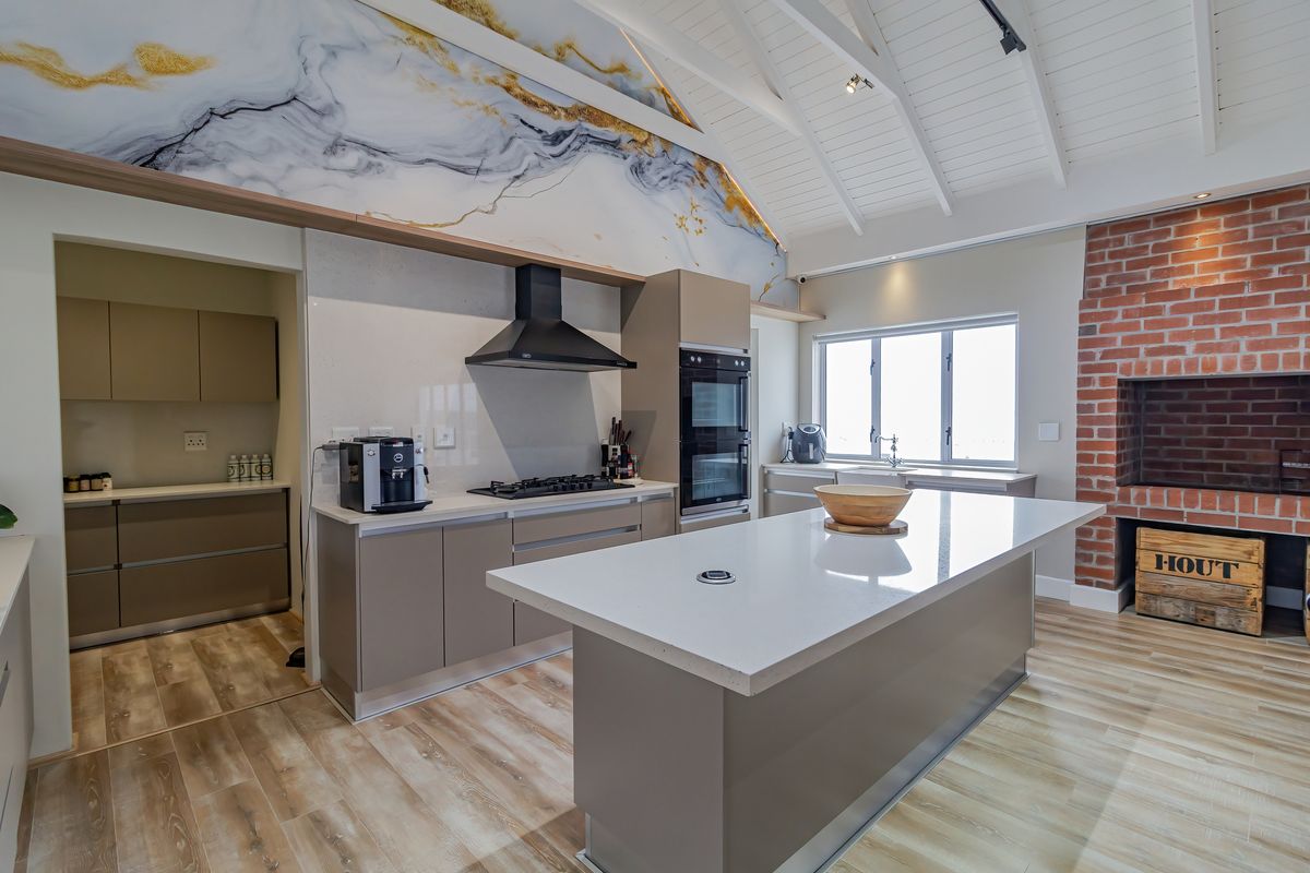 Modern kitchen featuring a central island, quartz countertops, built-in double oven, gas cooktop, and an adjacent scullery, complemented by a striking exposed brick fireplace.