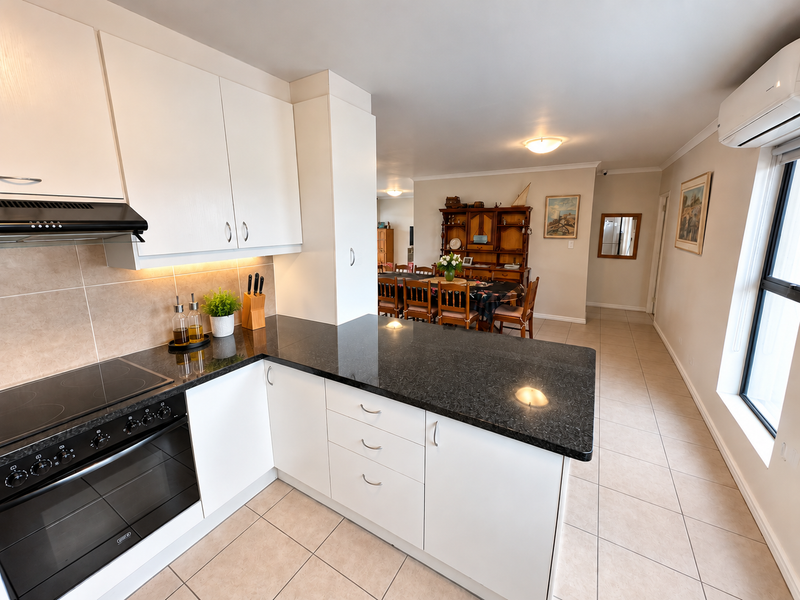 Open-plan kitchen and dining area featuring modern white cabinetry, dark granite countertops, integrated appliances, and tiled flooring throughout.