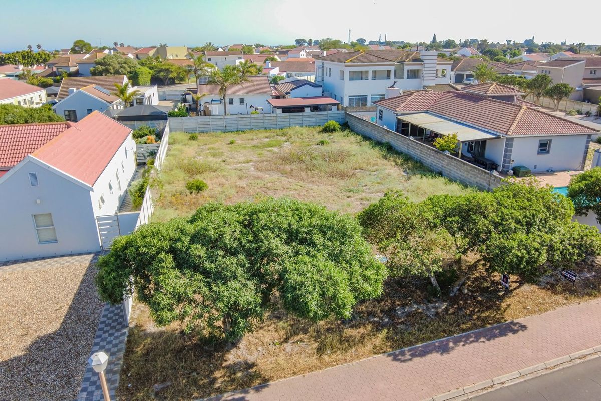 An aerial view of a vacant residential plot situated between existing homes, featuring established trees and boundary walls in a developed suburban neighborhood.
