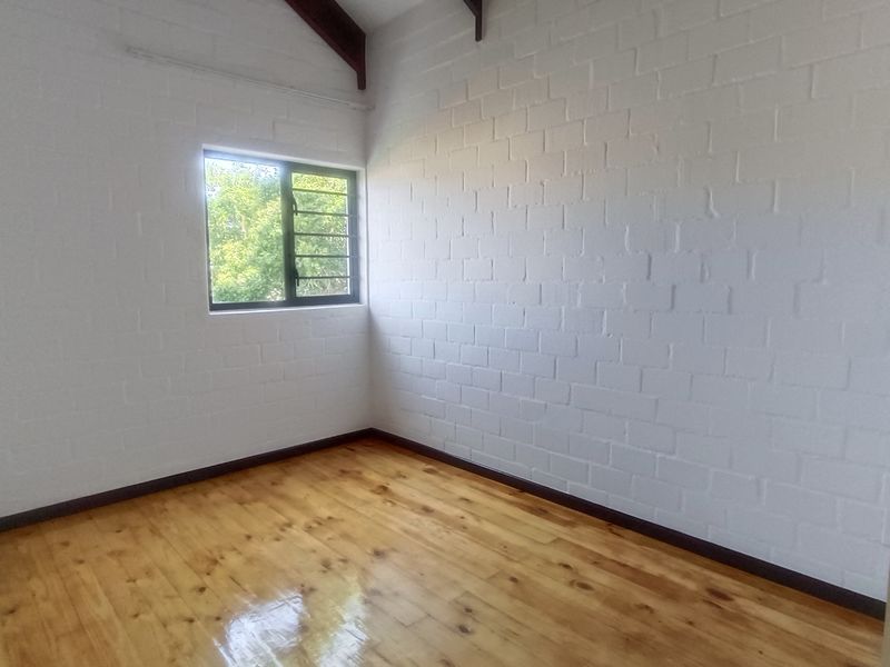A bright, empty bedroom featuring polished timber flooring, painted brick walls, and exposed timber rafters with a view through a louvre window.