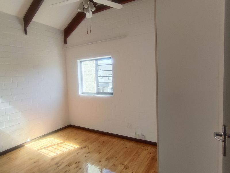 A bright, empty bedroom featuring vaulted ceilings with exposed timber beams, polished timber flooring, and painted brick walls.