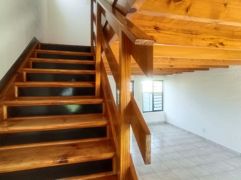 An open-plan interior space featuring a prominent timber staircase with balustrade, exposed wooden ceiling beams, tiled flooring, and painted brick walls.