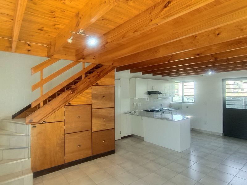 Open-plan living area featuring exposed timber ceiling beams, a wooden staircase with integrated storage, and a functional kitchen with a central island.