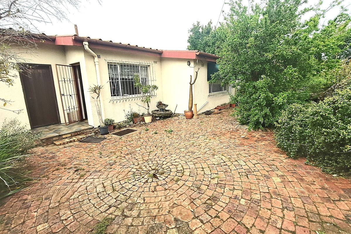 A rear exterior view featuring a circular brick paved patio area, security-fitted windows and doors, and established garden greenery.