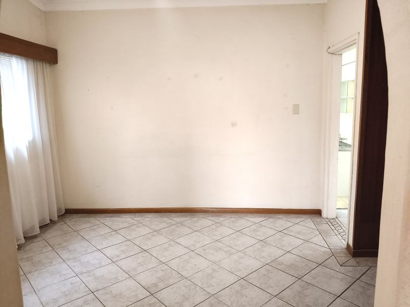 An empty living area featuring neutral tiled flooring, timber skirting boards, and a window pelmet, leading through to an adjacent kitchen space.