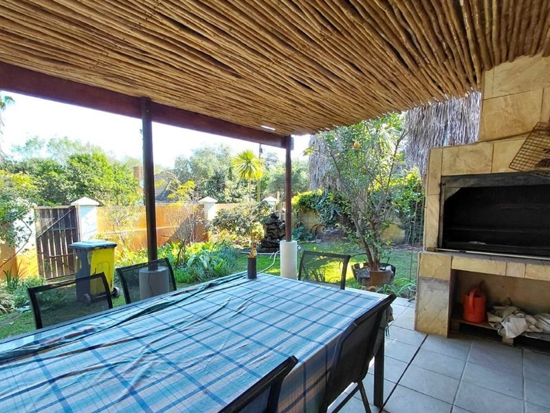 Covered outdoor patio featuring a rustic thatched roof, tiled flooring, and a built-in braai, overlooking a private garden area.