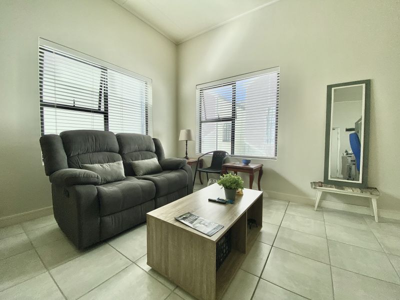 A bright living room featuring large windows with venetian blinds and neutral tiled flooring, offering a clean and functional space.