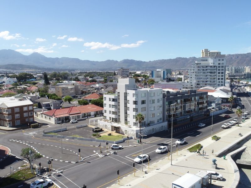 Aerial view of the apartment building situated in a prime coastal location, showcasing proximity to the beach, local roads, and surrounding urban landscape.