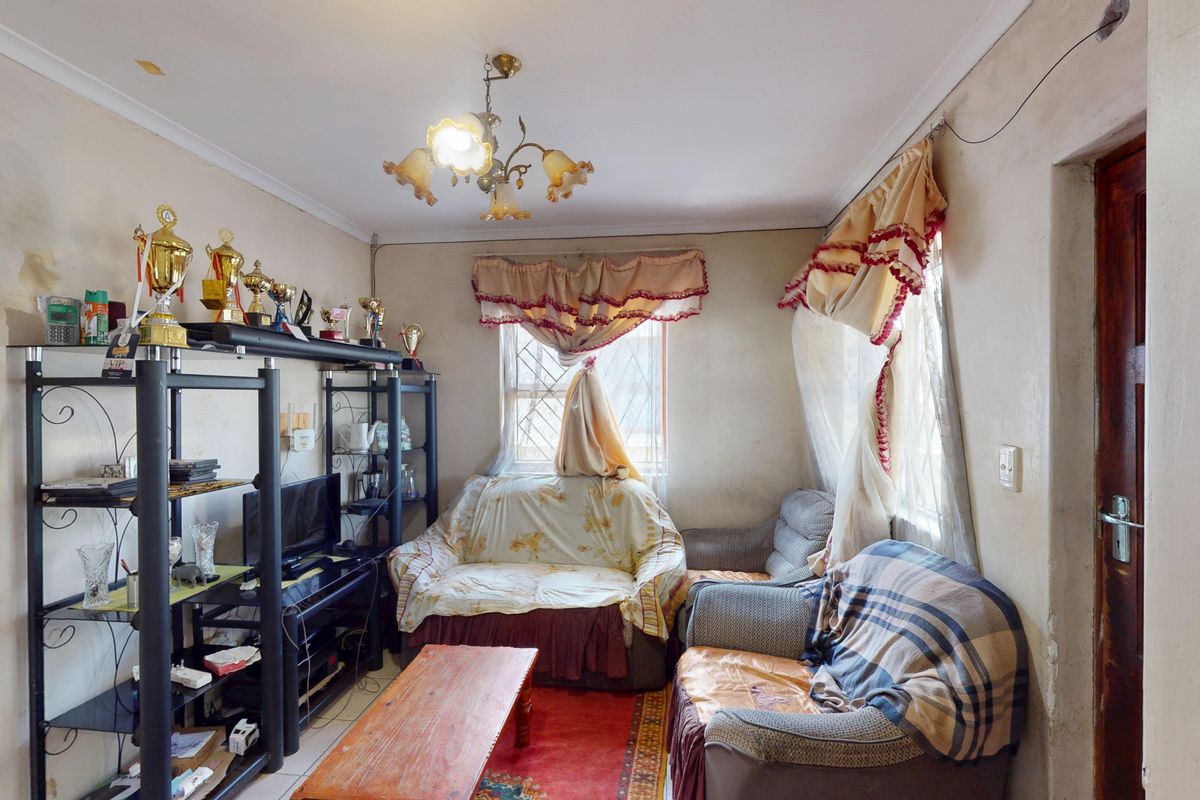 A living room area featuring tiled flooring, a central light fixture, and windows with security bars, currently requiring cosmetic updates and wall repairs.