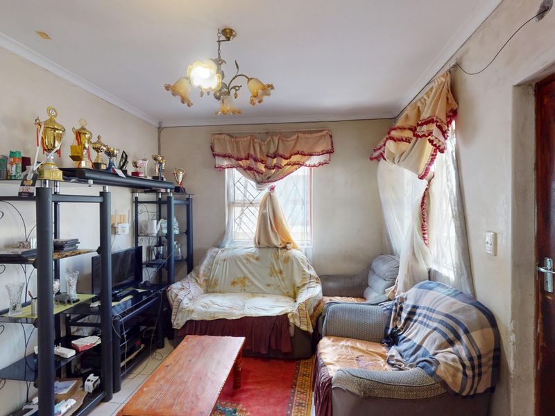 A living room area featuring tiled flooring, a central light fixture, and windows with security bars, currently requiring cosmetic updates and wall repairs.
