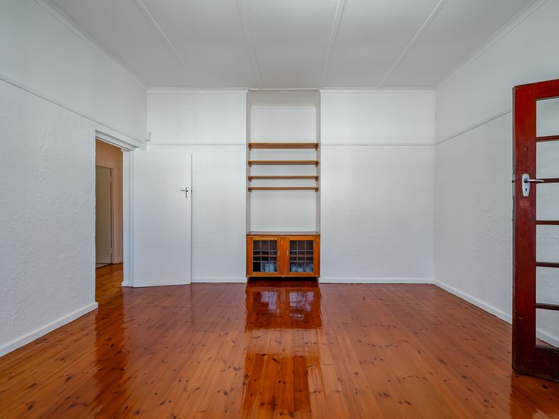 A bright, empty living room featuring polished timber floors, white walls with picture rails, and a built-in wooden cabinet with shelving.