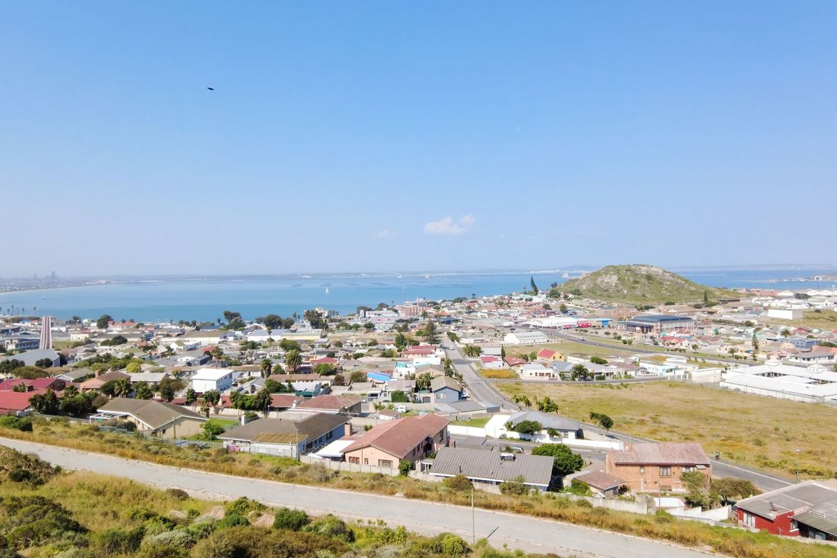 An elevated aerial view overlooking a residential coastal town, showcasing the surrounding landscape, road infrastructure, and proximity to the bay.