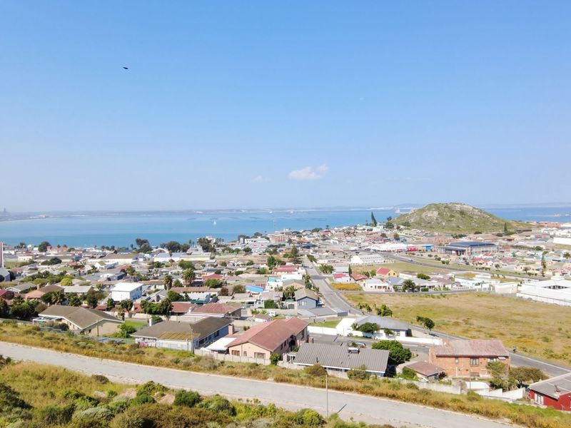 An elevated aerial view overlooking a residential coastal town, showcasing the surrounding landscape, road infrastructure, and proximity to the bay.