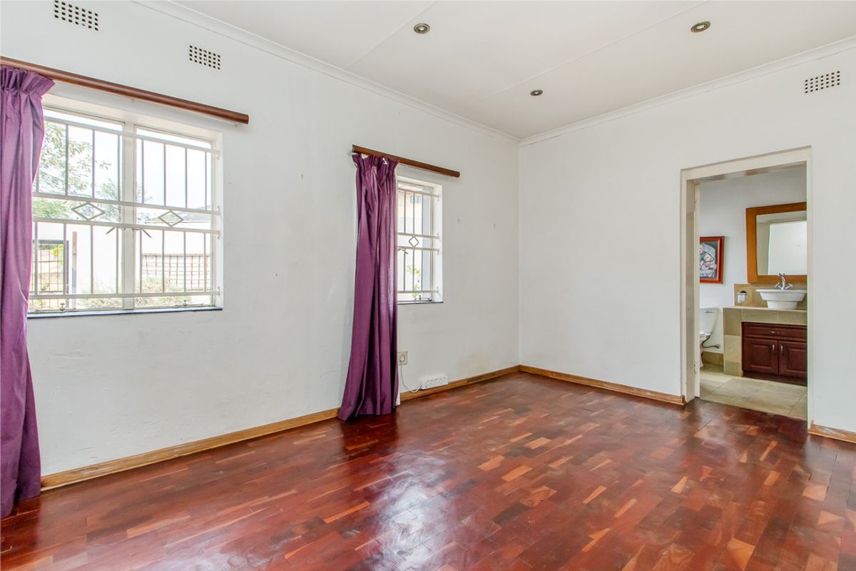 A spacious bedroom featuring polished parquet flooring, security bars on windows, and an adjoining en-suite bathroom with a vanity and toilet.