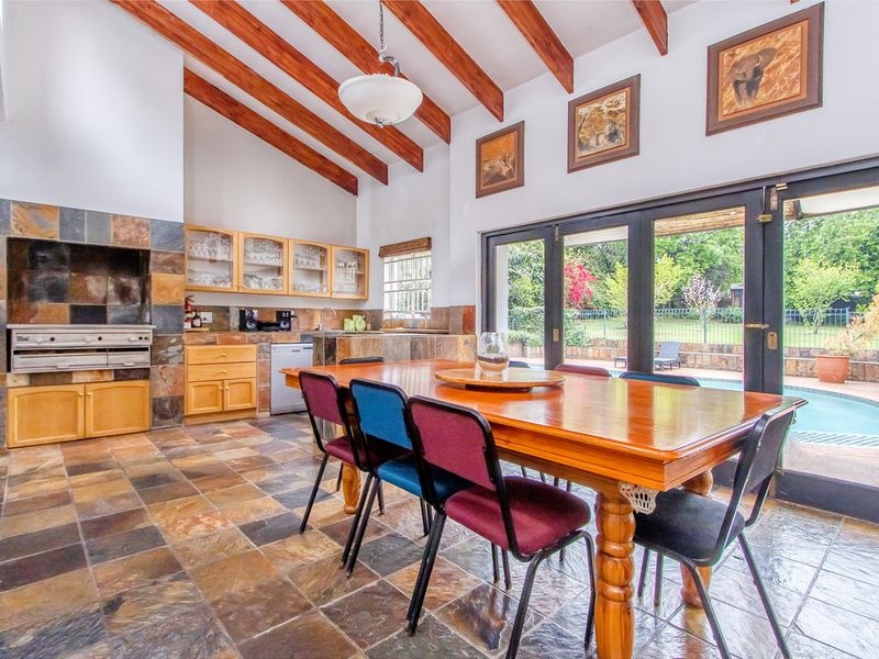 Spacious open-plan dining and entertainment area featuring exposed timber beams, slate flooring, a built-in braai, and bi-fold doors opening to the pool area.