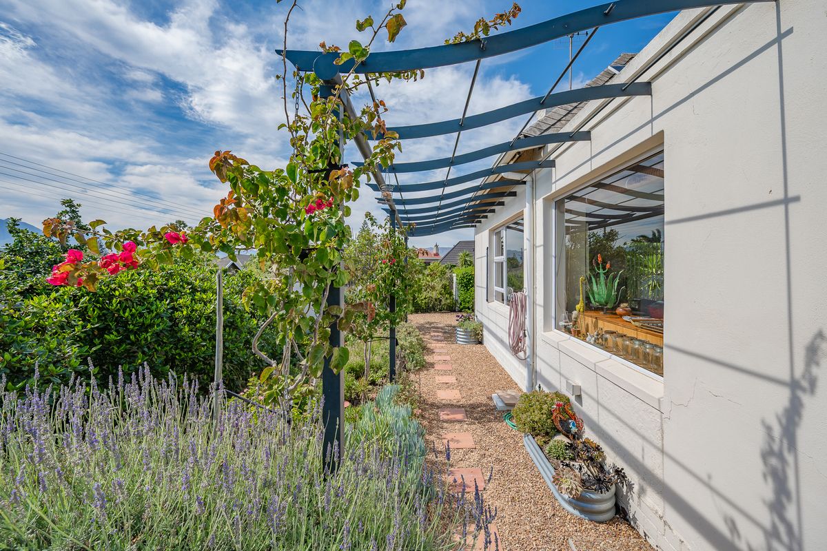 A well-maintained rear exterior featuring a metal pergola, gravel pathway, and established garden beds alongside the home's exterior wall.