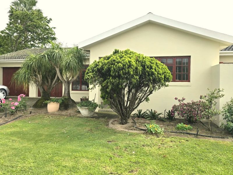 Front elevation of a single-story house featuring a manicured lawn, established garden beds, and a paved driveway leading to the garage.