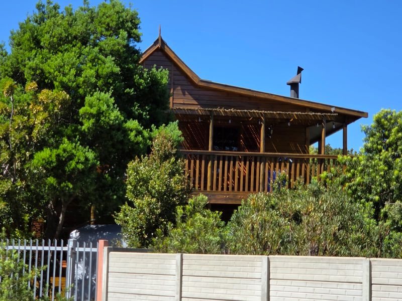 A charming timber-clad house featuring a raised wooden balcony, chimney, and secure perimeter fencing, set within a lush, established garden.