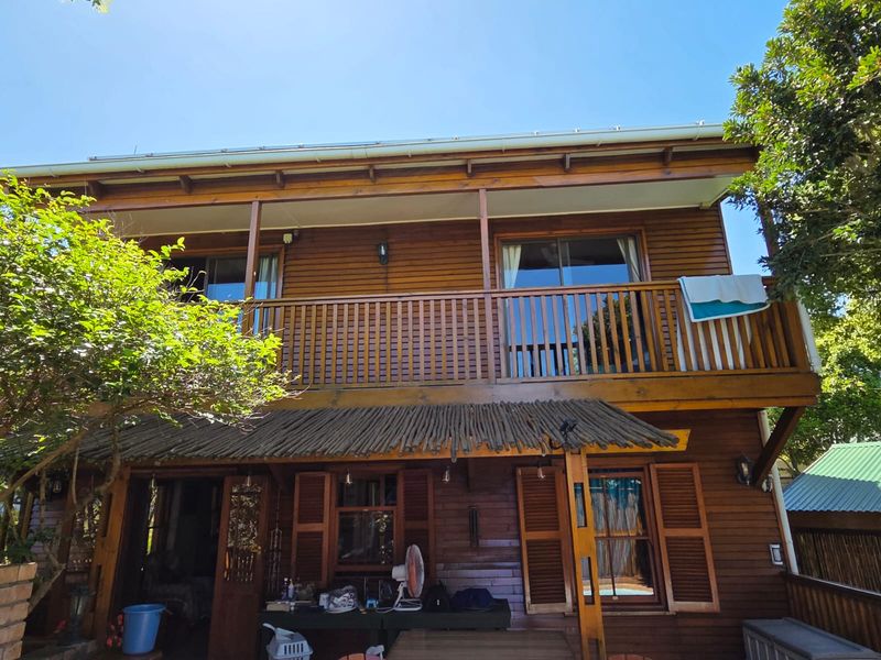A two-story timber-clad home featuring a spacious upper-level balcony with timber balustrades and a charming thatched pergola over the lower patio area.