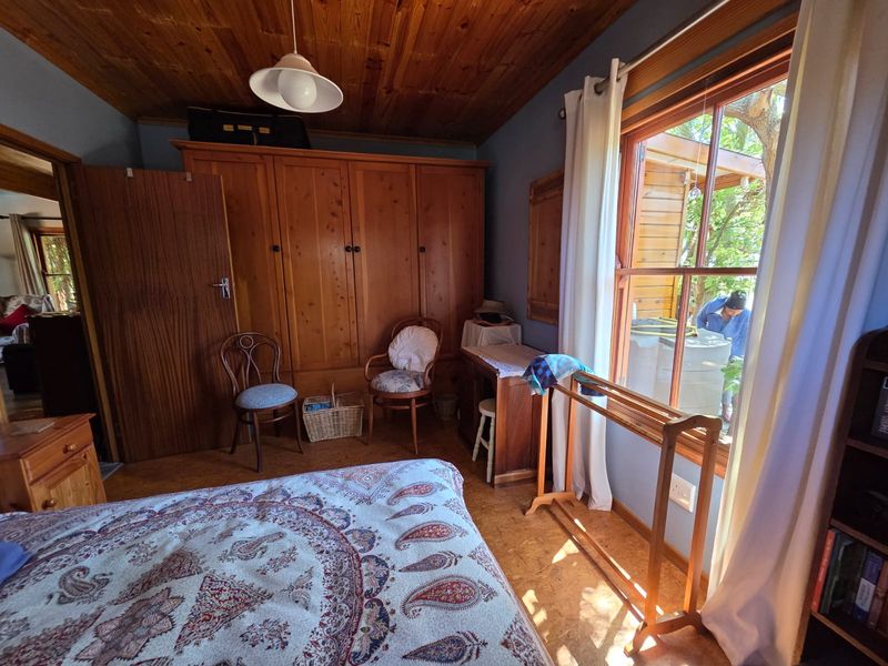 A cozy bedroom featuring a timber ceiling, cork flooring, and a large built-in wooden wardrobe, with natural light entering through a sash window.