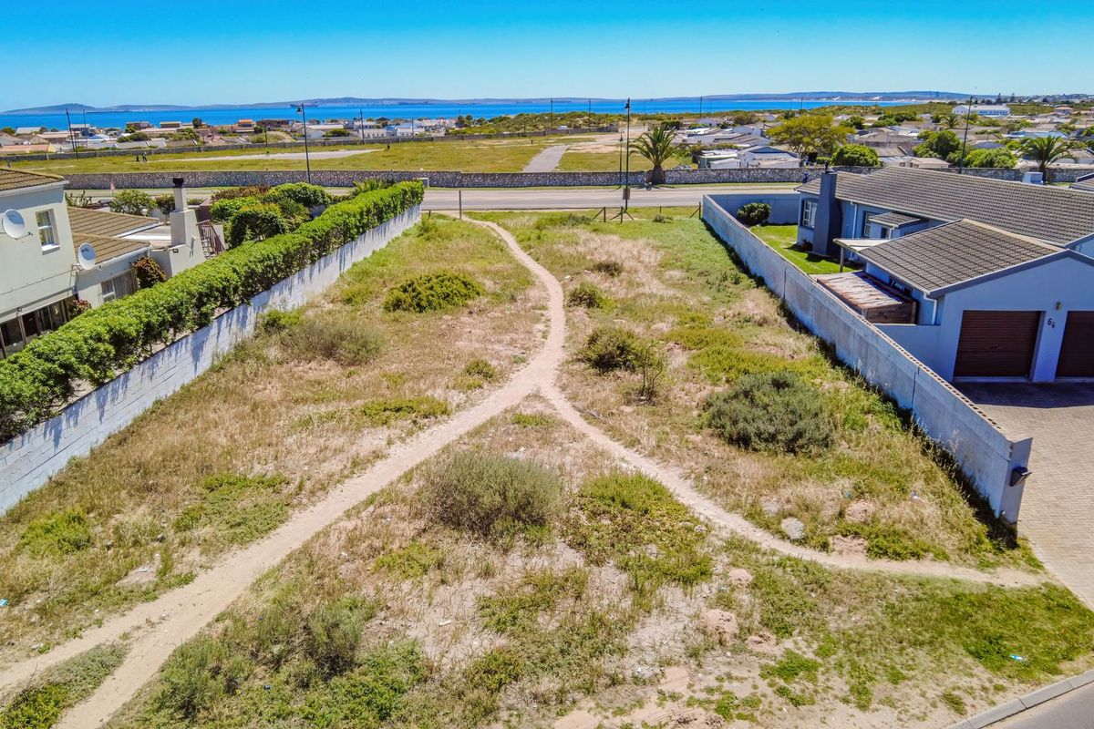 Aerial view of a vacant residential plot situated between existing homes, featuring boundary walls and a clear path, with distant coastal views.
