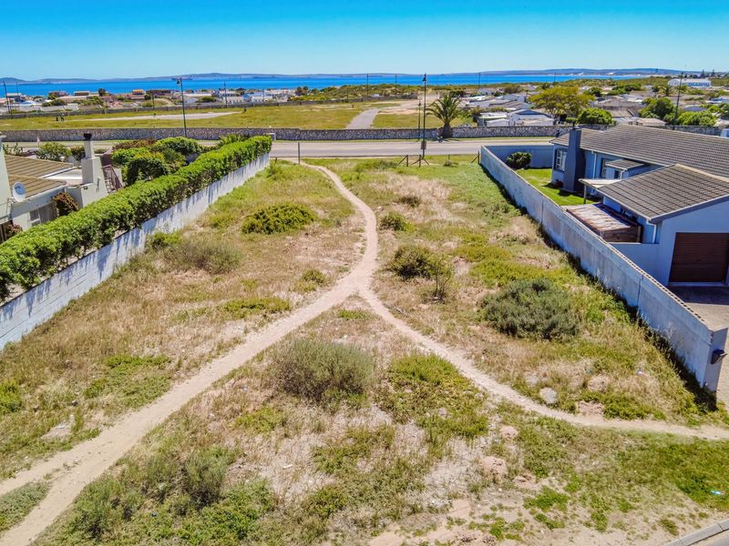 Aerial view of a vacant residential plot situated between existing homes, featuring boundary walls and a clear path, with distant coastal views.