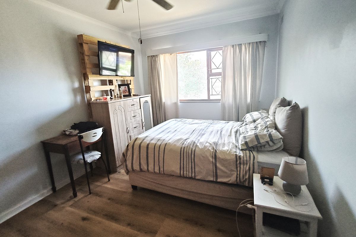 A cozy bedroom featuring laminate flooring, a ceiling fan, and a window with security bars, offering a comfortable and functional living space.