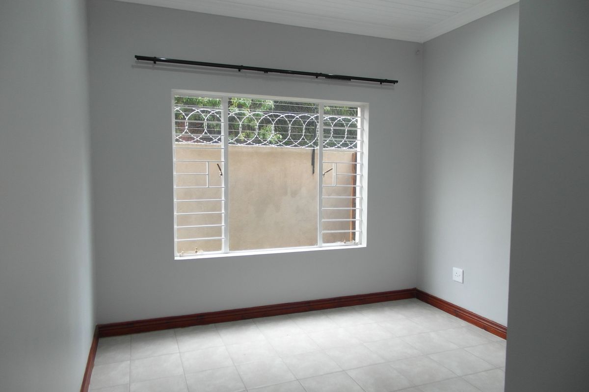 A clean, empty bedroom featuring neutral grey walls, tiled flooring, timber skirting, and a window fitted with security bars.
