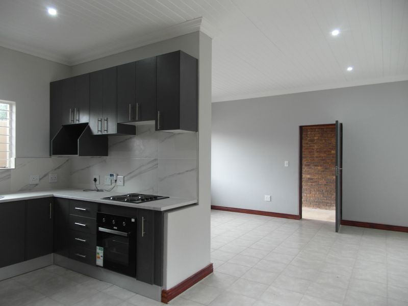 Modern open-plan kitchen featuring dark cabinetry, built-in oven and hob, marble-look splashback, and neutral tiled flooring, leading into an adjacent living area.